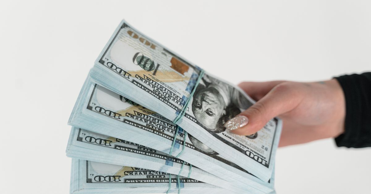 Close-up of a hand holding several stacks of US hundred dollar bills on a white background.